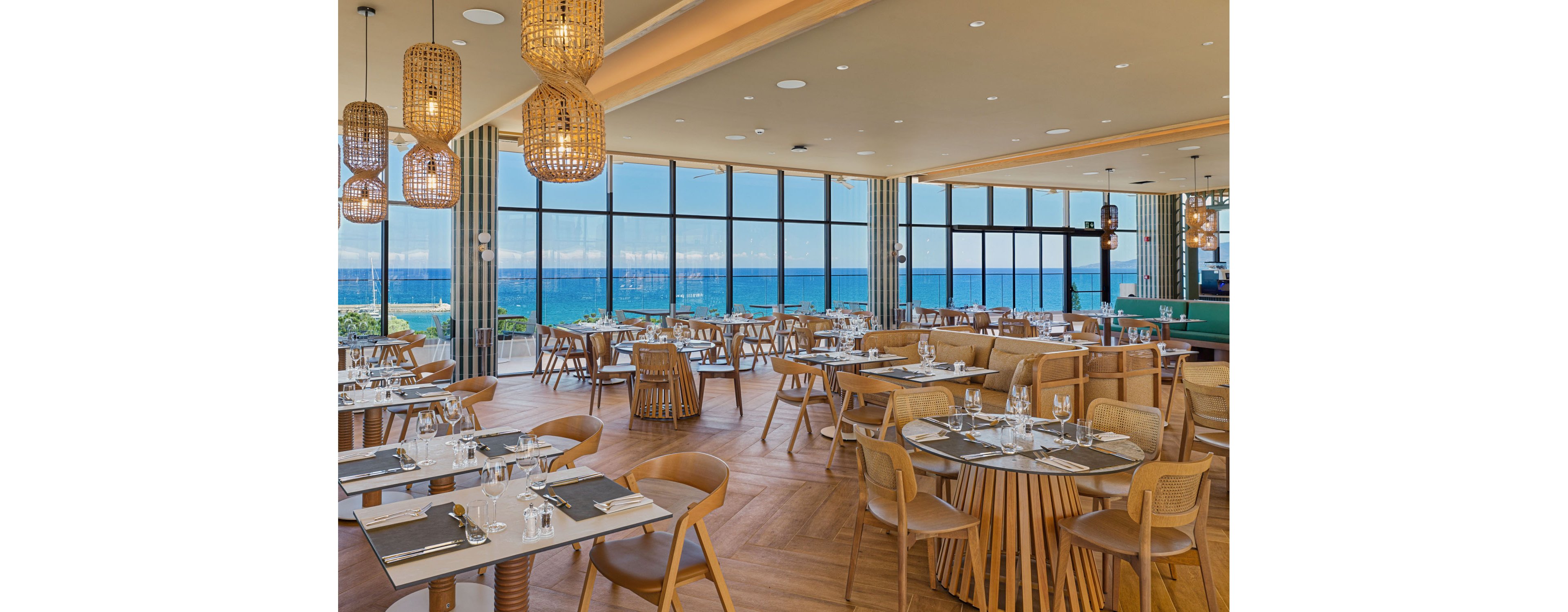 A contemporary restaurant dining area features neatly arranged wooden tables and chairs. Floor-to-ceiling windows reveal a bright blue ocean and clear sky outside. The interior design includes warm wood tones, woven pendant lights, and minimalist table settings with glassware and cutlery. The space appears calm and inviting, ideal for relaxed seaside dining.