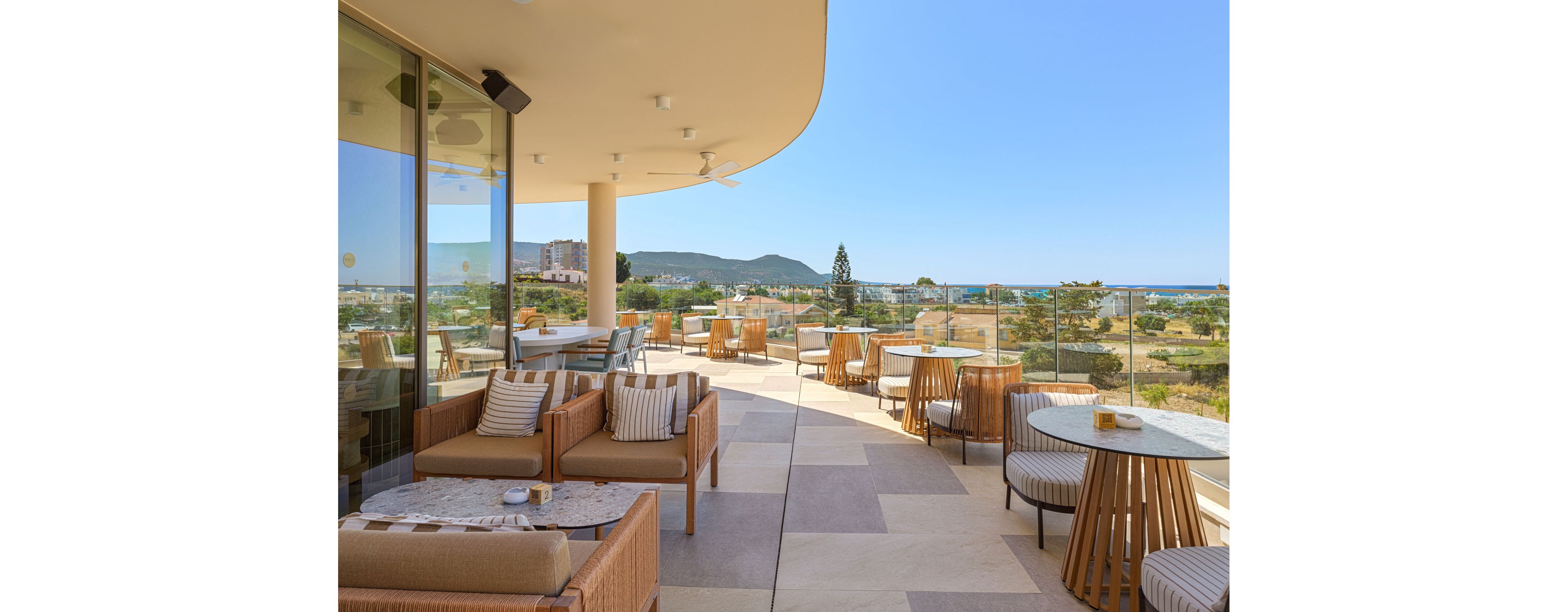 A contemporary outdoor hotel terrace features cushioned lounge seating and round glass-top tables. The space overlooks a coastal landscape with distant hills and blue sea under a clear sky. Neutral-toned furniture and striped pillows create a relaxed, modern atmosphere. Large glass railings and curved architecture enhance the open, airy feel of the setting.