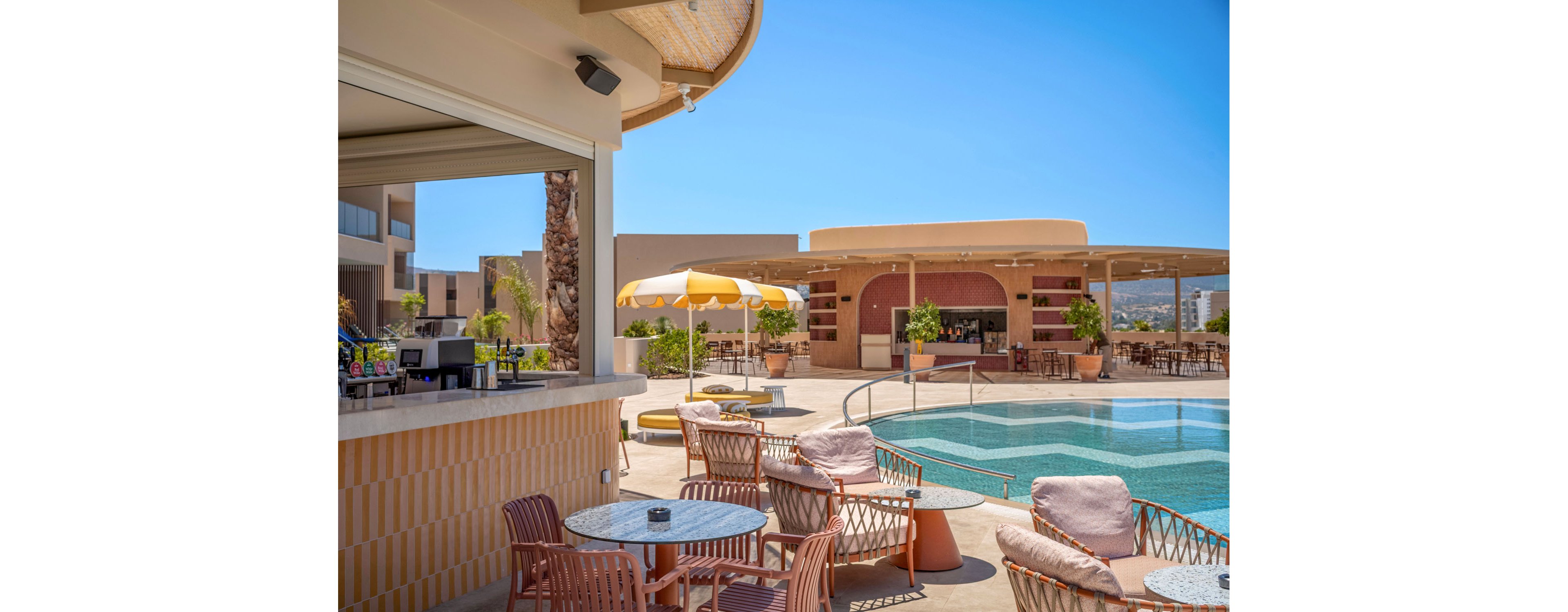 A contemporary resort pool area features a stylish open-air bar and comfortable lounge seating. Pink and beige chairs with cushions surround small tables near the pool, creating a relaxed social setting. A yellow-and-white striped umbrella provides shade beside the water, while the bar pavilion and surrounding buildings frame the scene. Clear blue skies and distant hills emphasize a sunny, vacation atmosphere.