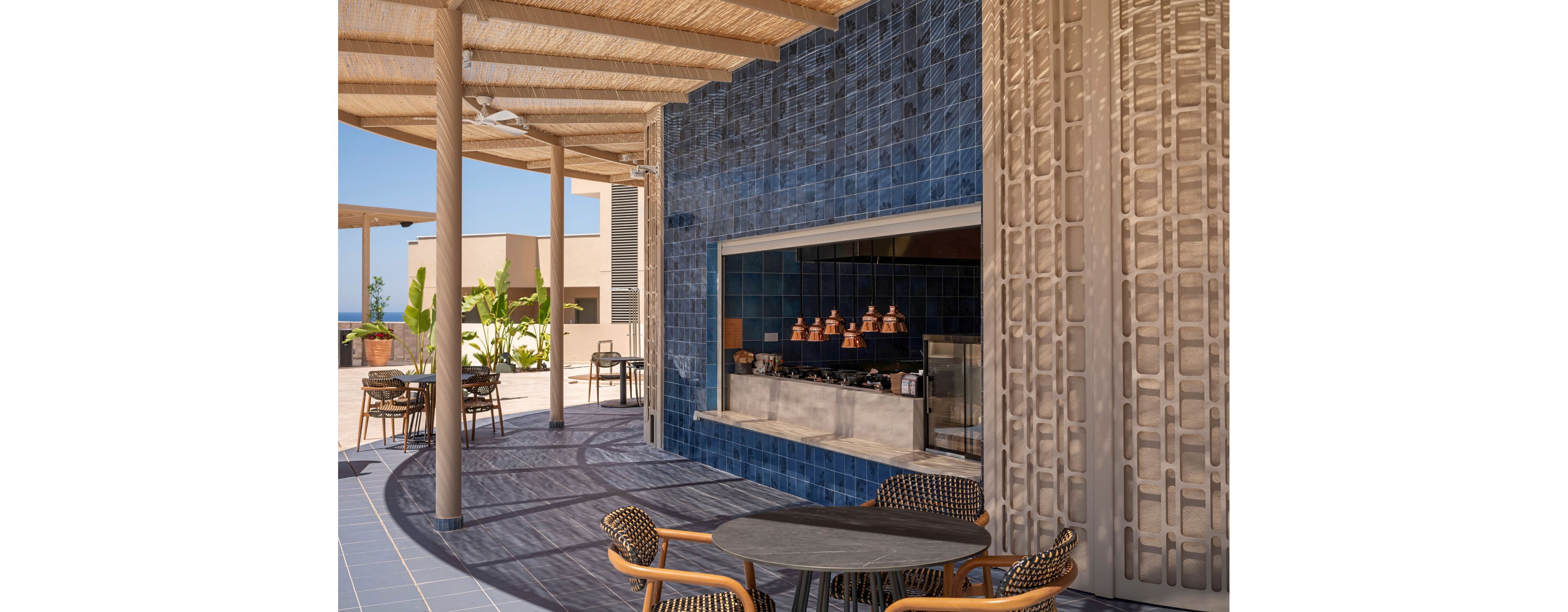 An open-air cafe features a modern serving counter framed by deep blue tiles. Copper pots and kitchen equipment are visible inside the recessed cooking area. The seating area includes round tables and woven chairs arranged under a wooden pergola that casts patterned shadows on the floor. In the background, additional tables, potted plants, and contemporary buildings create a relaxed resort-like atmosphere.