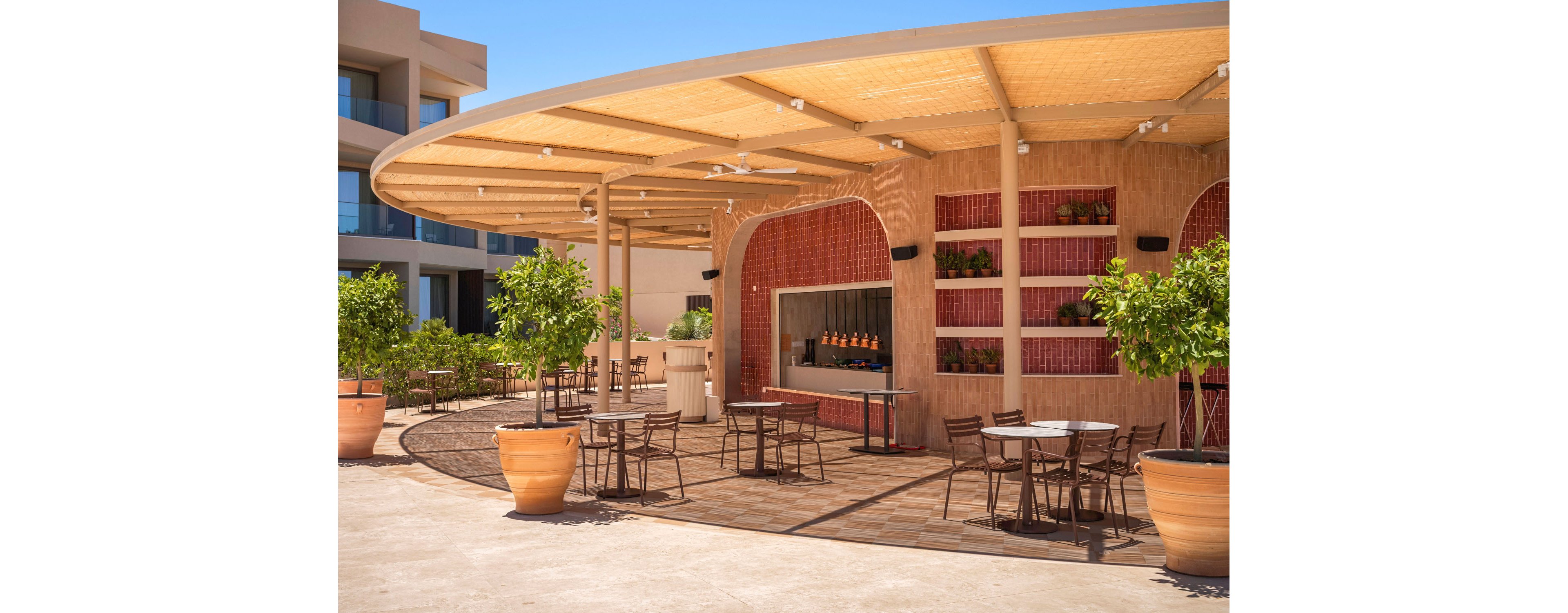 An open-air bar with a curved, shaded roof sits in a sunny resort courtyard. Several small tables and metal chairs are arranged on a wooden deck, creating a relaxed seating area. Large terracotta planters with green trees frame the space, while shelves behind the bar display bottles and potted plants. Modern resort buildings and a clear blue sky complete the bright, vacation-like atmosphere.