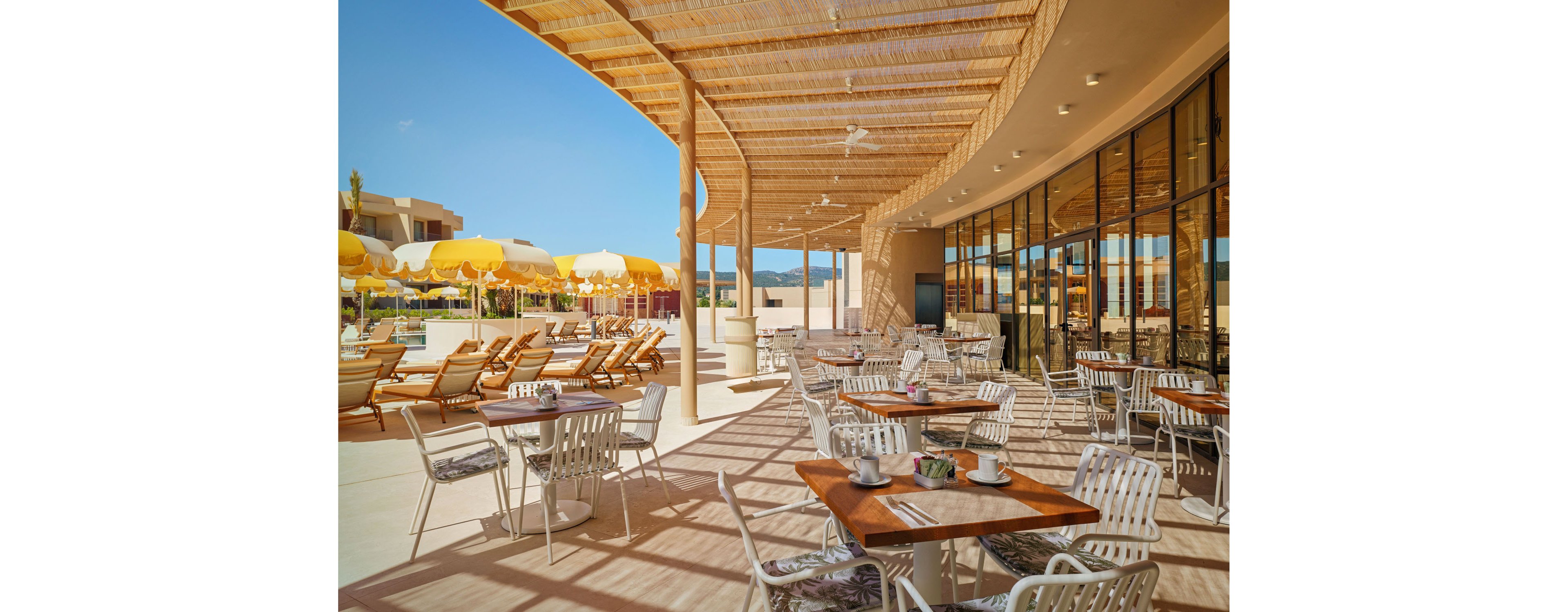 An outdoor resort café features neatly arranged tables and chairs under a woven pergola. Beyond the terrace, rows of sun loungers and bright yellow umbrellas line a poolside deck. The scene is bathed in warm sunlight with clear blue skies and distant hills in the background. Reflections in the large glass windows add depth and emphasize the modern, relaxed vacation atmosphere.