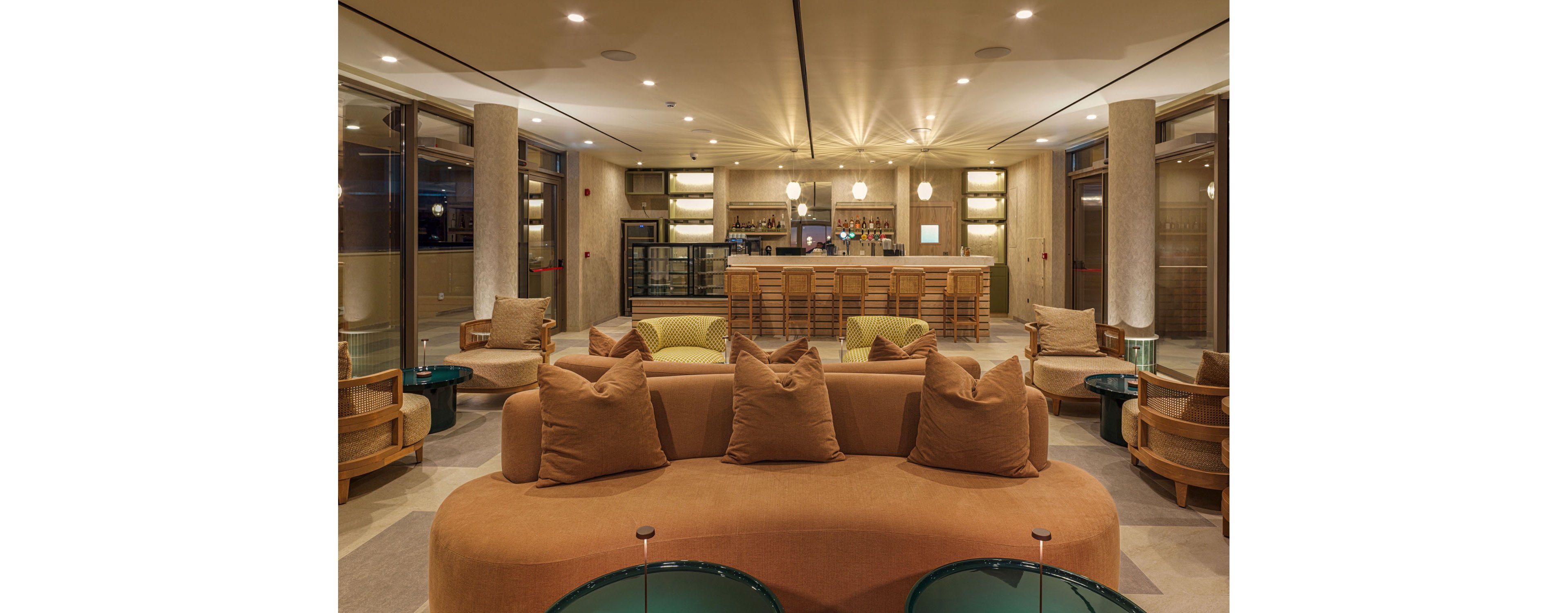A contemporary hotel lounge features a curved sofa with large brown cushions in the foreground. Matching armchairs and low glass tables are arranged to create an inviting seating area. In the background, a stylish bar with wooden stools and illuminated pendant lights adds a focal point. Floor-to-ceiling windows and neutral tones contribute to a warm, sophisticated atmosphere.