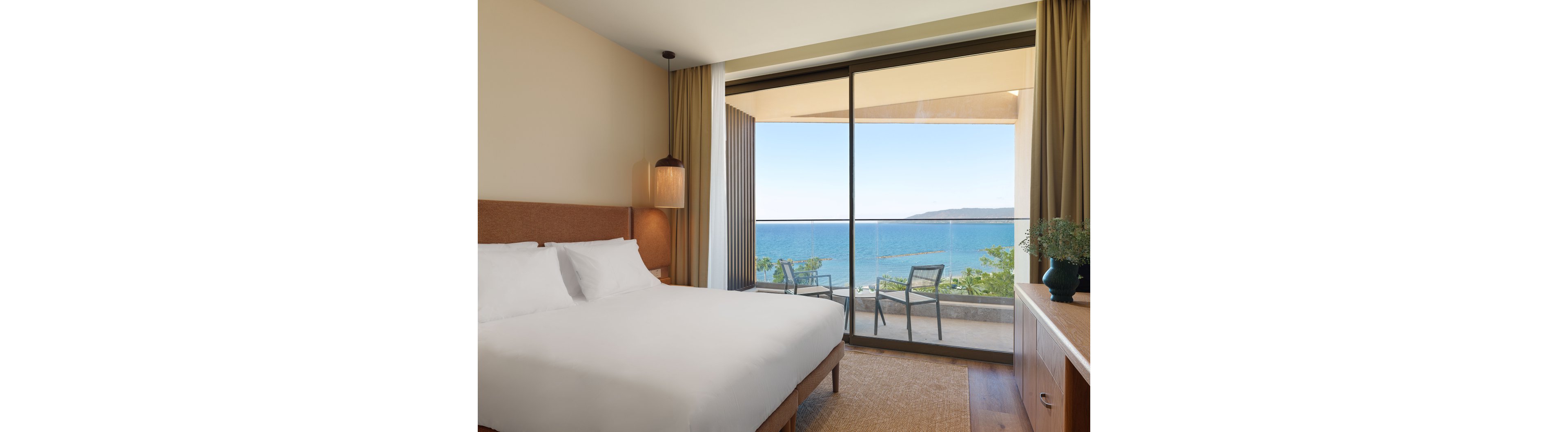 A contemporary hotel bedroom features a large neatly made bed with white linens and warm neutral tones. Floor-to-ceiling sliding glass doors open to a balcony furnished with two chairs and a small table. Beyond the balcony, a calm blue ocean and distant coastline are visible under a clear sky. Soft lighting, simple decor, and natural materials create a relaxing, minimalist atmosphere.