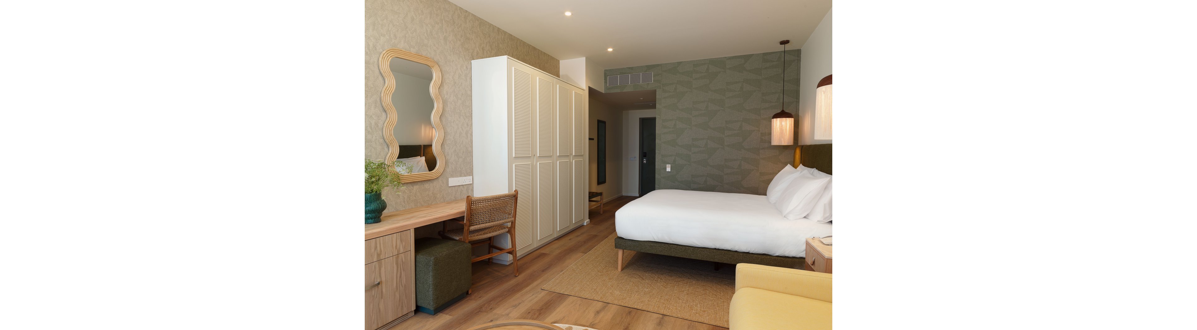A contemporary hotel bedroom features a large neatly made bed with white linens and a green upholstered base. The room includes a wooden desk with a wicker chair, a small ottoman, and a light wood wardrobe along one wall. Soft pendant lighting, patterned accent wallpaper, and a decorative mirror add warmth and style to the space. The overall atmosphere is calm and inviting, with neutral tones and natural textures creating a modern, minimalist look.