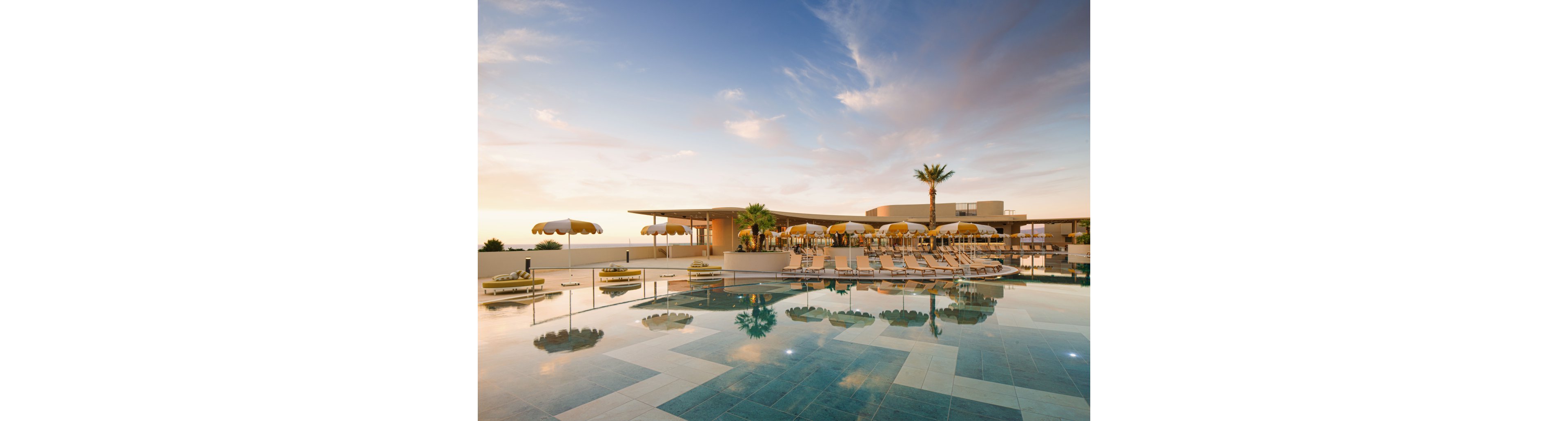 A serene rooftop swimming pool reflects the pastel colors of a sunset sky. Lounge chairs and yellow umbrellas line the deck, creating a relaxed resort atmosphere. A single palm tree stands near the seating area, adding a tropical touch to the modern architecture. The calm water and soft lighting emphasize a tranquil, luxurious vacation setting.