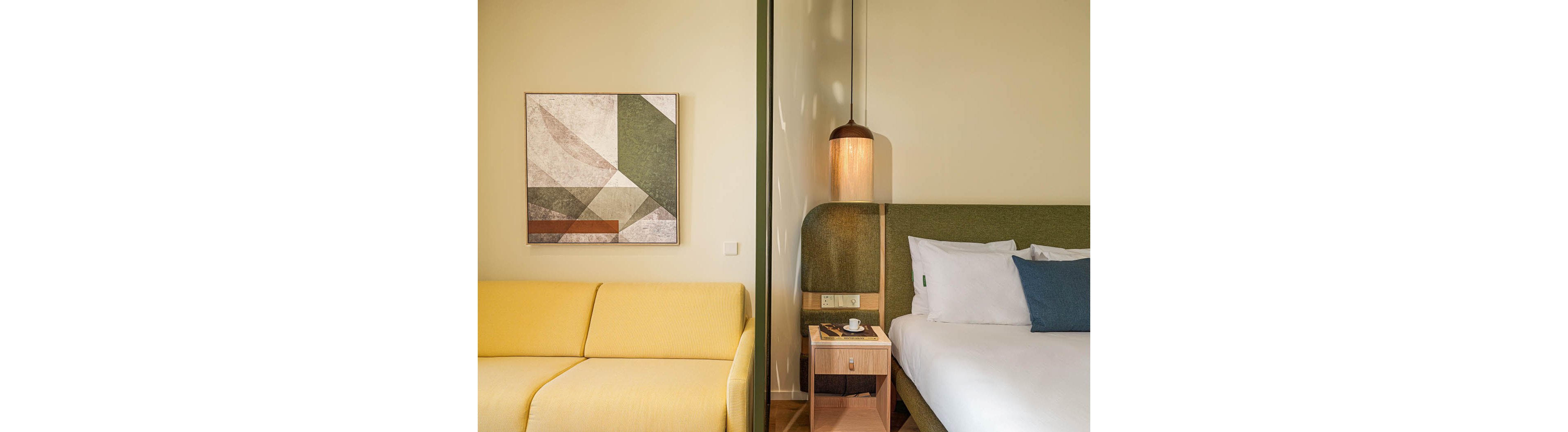 A contemporary hotel room features a neatly made bed with white linens and a blue accent pillow. Beside the bed, a wooden nightstand holds a cup and saucer under a warm hanging lamp. A yellow sofa sits on the opposite side of a partial divider wall, adding a bright contrast to the space. Geometric artwork in muted tones decorates the wall, creating a stylish and cozy atmosphere.