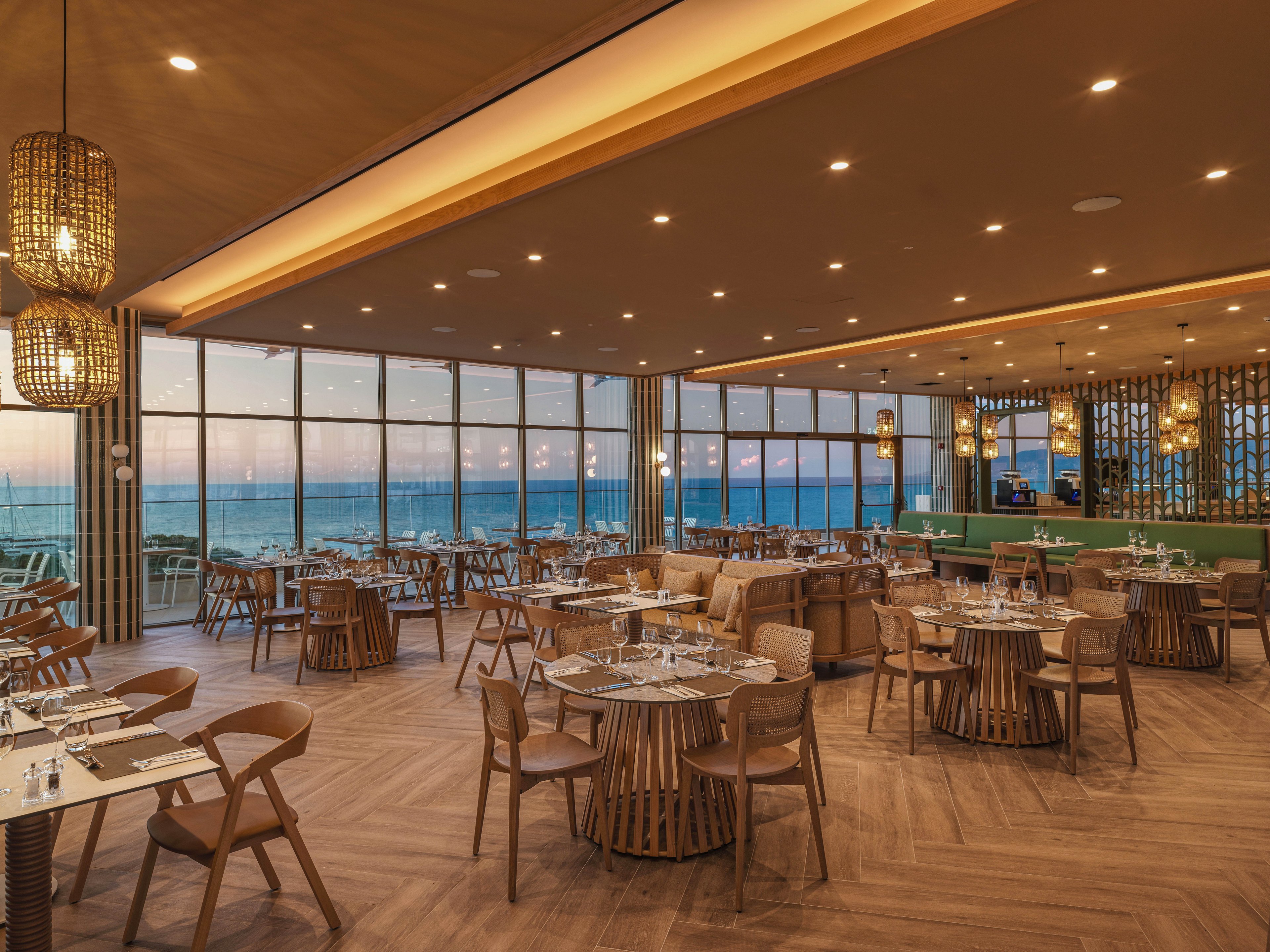 A contemporary restaurant dining room overlooks the ocean through floor-to-ceiling windows at sunset. Round wooden tables and matching chairs are neatly arranged with glassware and place settings. Warm ceiling lights and woven pendant lamps create a cozy, inviting atmosphere. The space features a wooden floor, soft neutral tones, and a bar area in the background.