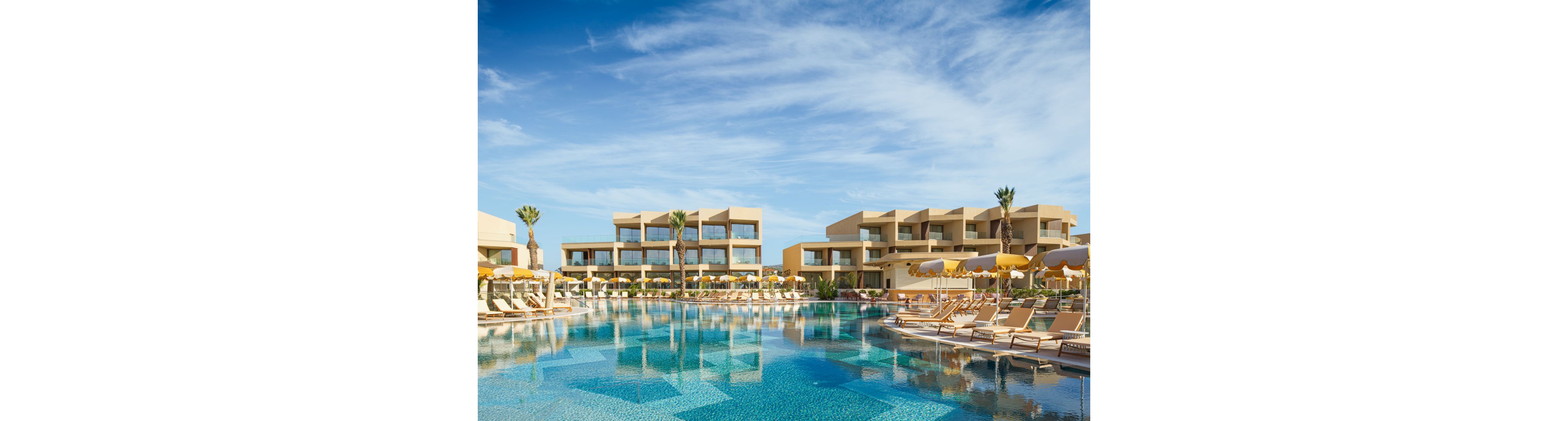 A contemporary resort complex features multiple low-rise beige buildings surrounding a large, clear blue swimming pool. Numerous sun loungers and yellow umbrellas line the poolside, suggesting a relaxing vacation setting. Tall palm trees and a bright blue sky with light clouds enhance the tropical, sunny atmosphere. The calm water reflects the architecture and sky, emphasizing tranquility and leisure.