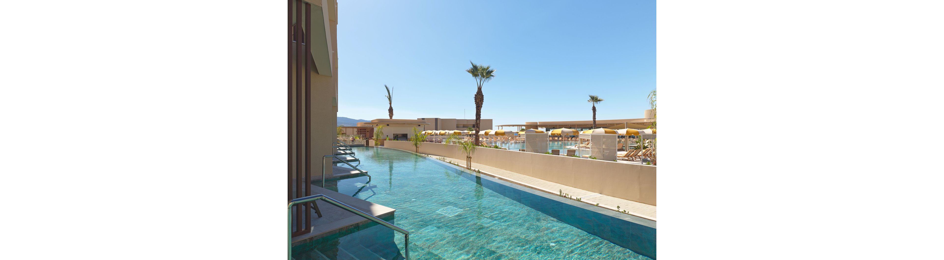 A sleek, modern swimming pool stretches alongside a resort building under a clear blue sky. In the background, guests relax on sun loungers shaded by yellow and white umbrellas. Palm trees and desert mountains frame the scene, emphasizing a warm, sunny climate. The calm turquoise water and clean architectural lines create a tranquil, luxurious atmosphere.