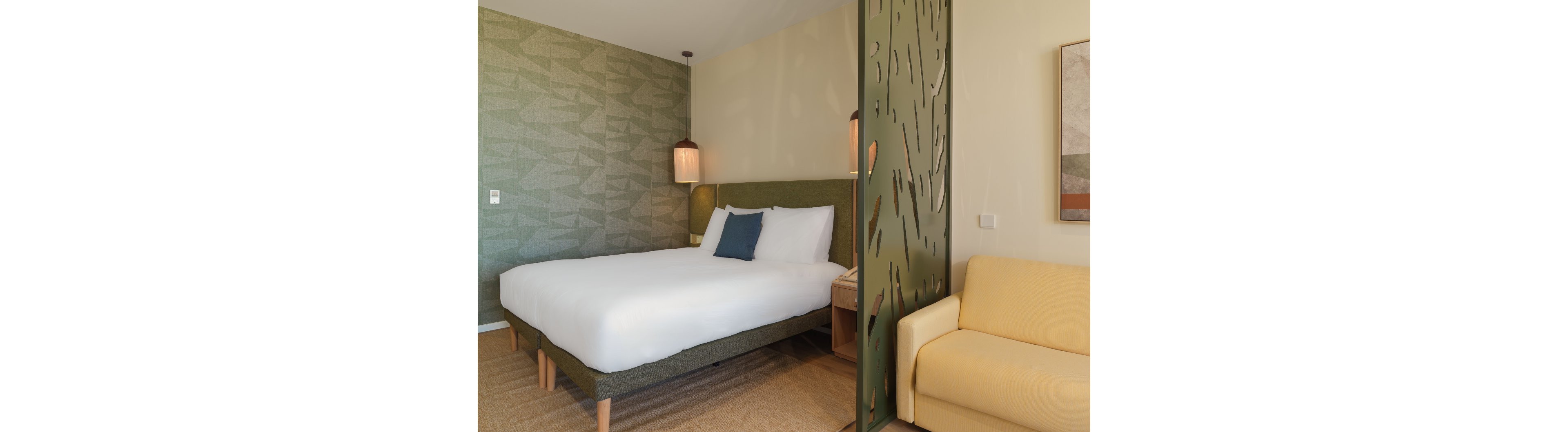 A contemporary hotel room features a large neatly made bed with white linens and a green upholstered frame. A decorative green partition with cutout patterns separates the sleeping area from a light yellow sofa. Warm pendant lights hang above the bed, complementing the textured green accent wall. The overall design feels clean, minimal, and inviting, with neutral tones and subtle geometric details.