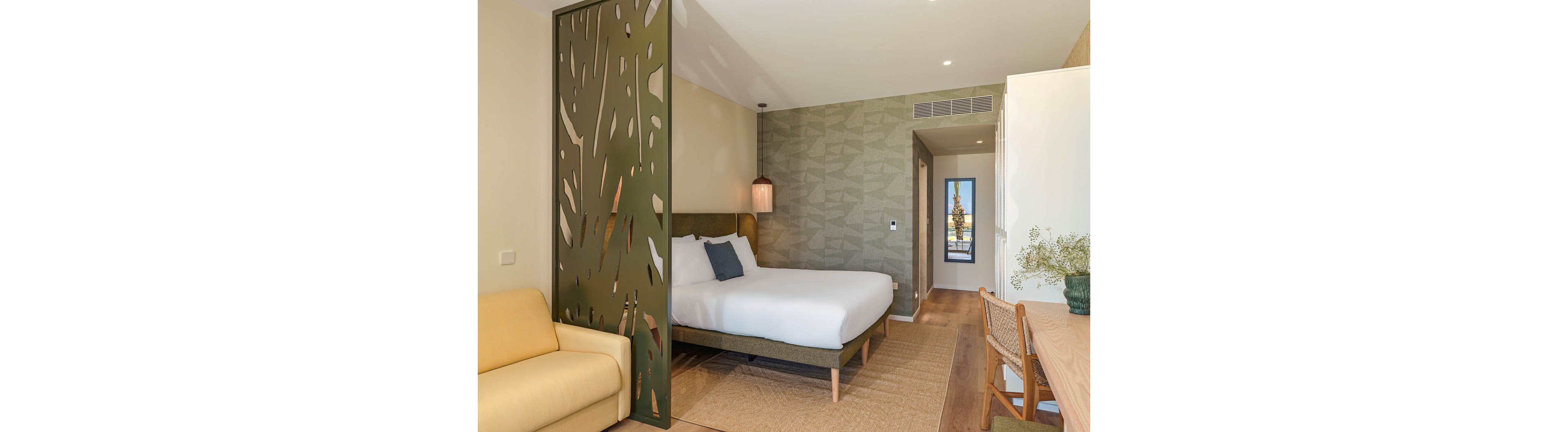 A contemporary hotel bedroom features a large bed with white linens and a green upholstered headboard. A decorative perforated partition separates the sleeping area from a small sitting space with a yellow sofa. The room includes a wooden desk with a woven chair, minimalist decor, and a patterned accent wall. Natural light enters through a doorway at the far end, creating a bright and inviting atmosphere.