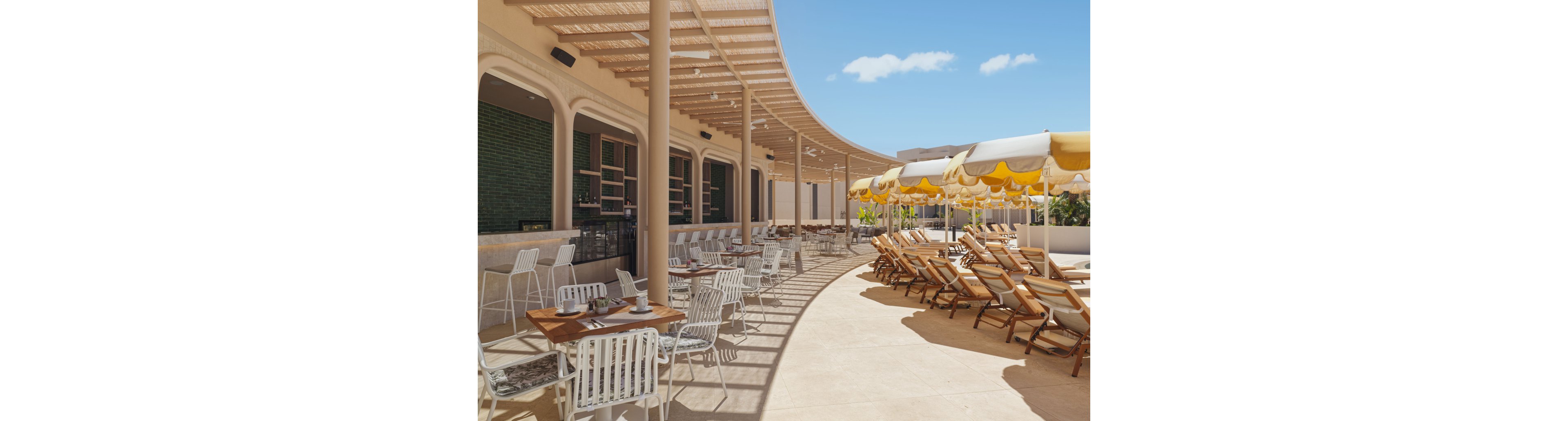An open-air cafe terrace features neatly arranged tables and white metal chairs under a curved pergola. A bar with green tiled walls lines one side, while rows of wooden sun loungers sit beneath yellow-and-white striped umbrellas. The bright midday light and clear blue sky create a warm, inviting atmosphere. The scene suggests a relaxed resort or beachside setting ideal for outdoor dining and lounging.