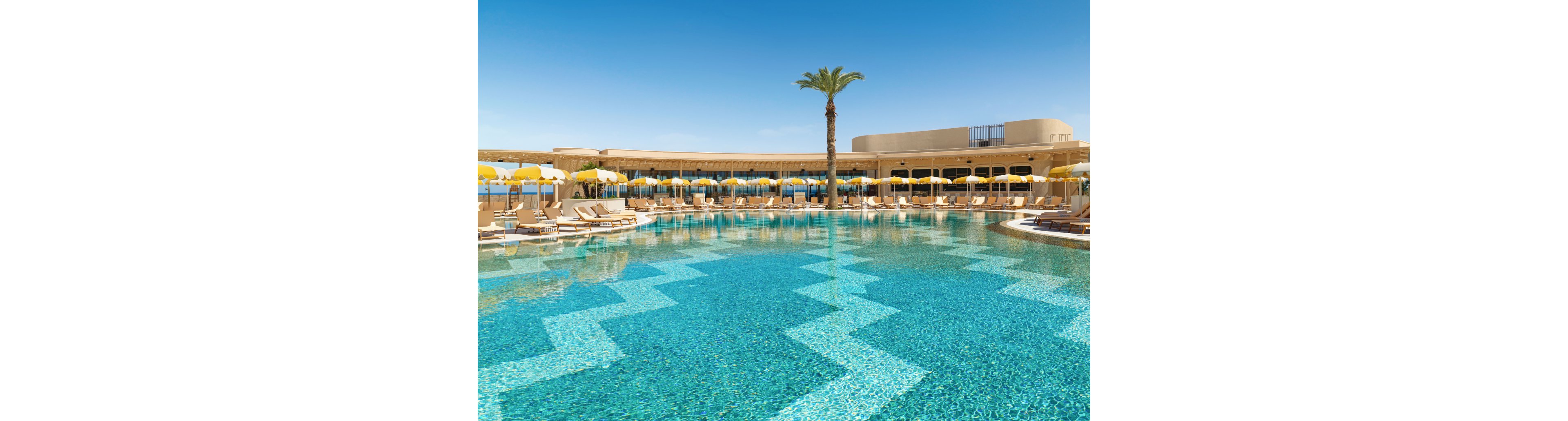 A large, clear blue swimming pool dominates the foreground of a sunny resort scene. Beige lounge chairs and yellow-and-white umbrellas line the curved poolside terrace, facing toward the water. A single tall palm tree rises near the center, adding a tropical touch against the bright blue sky. In the distance, the horizon suggests a coastal location with views toward the sea.