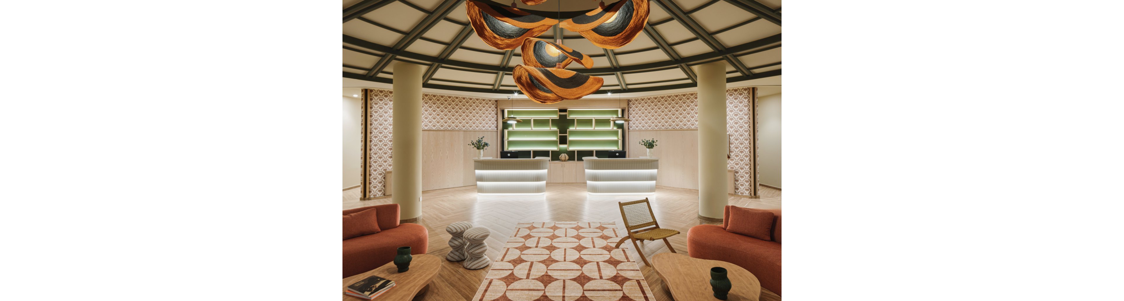 A contemporary hotel lobby features a central seating area with curved sofas and sculptural side tables. In the background, two illuminated reception desks stand against a wall with green accent panels and patterned wallpaper. A geometric rug and warm wood flooring complement the neutral and terracotta color palette. Overhead, artistic pendant lights hang from a coffered ceiling, creating a welcoming and sophisticated atmosphere.