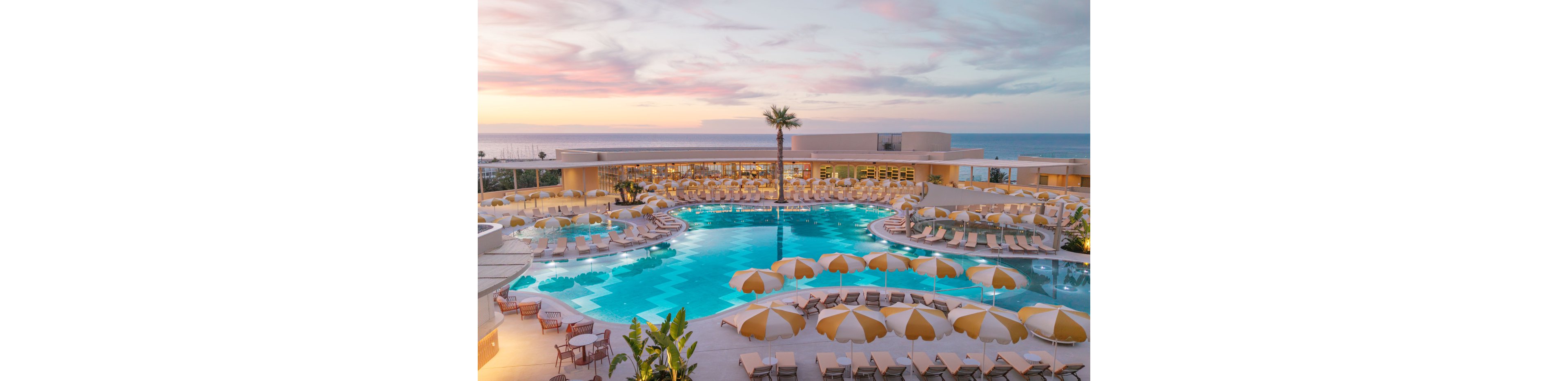 A large resort swimming pool curves around a central palm tree overlooking the ocean at sunset. Rows of yellow and white umbrellas and sun loungers surround the pool, creating a symmetrical and inviting layout. The sky is filled with soft pastel clouds that reflect in the calm blue water. Modern low-rise resort buildings frame the scene, emphasizing a relaxed coastal vacation atmosphere.