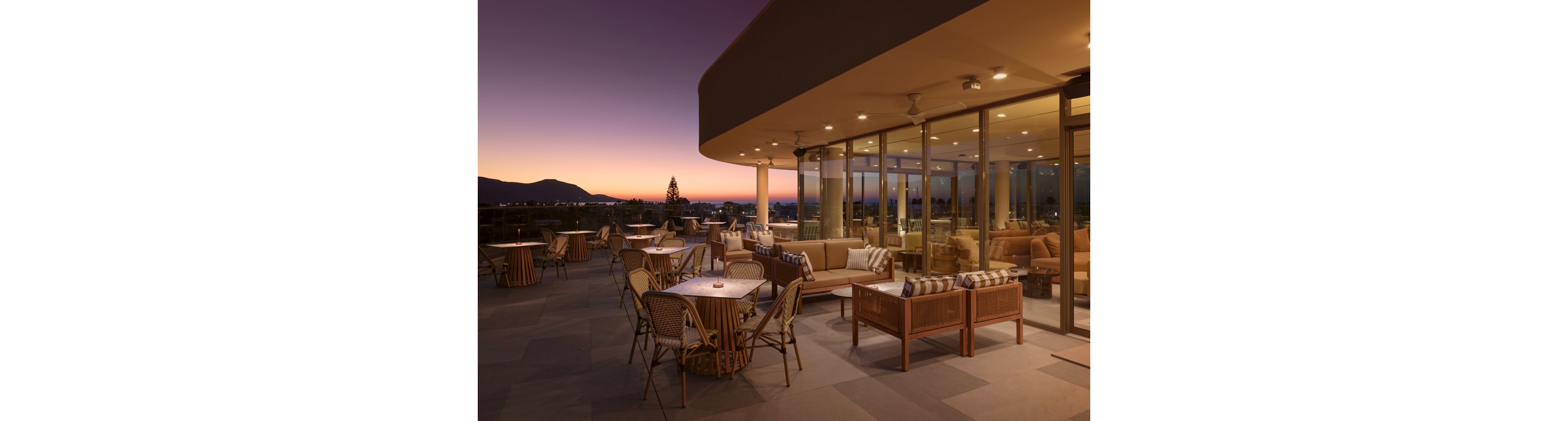 An elegant rooftop terrace lounge is shown at dusk with comfortable sofas and café-style tables arranged across a spacious patio. Floor-to-ceiling glass walls reveal a warmly lit indoor seating area, creating a seamless transition between interior and exterior spaces. The sky glows with a soft gradient from orange to deep purple, silhouetting distant mountains and city lights. The overall scene feels calm and inviting, ideal for evening dining or relaxed social gatherings.