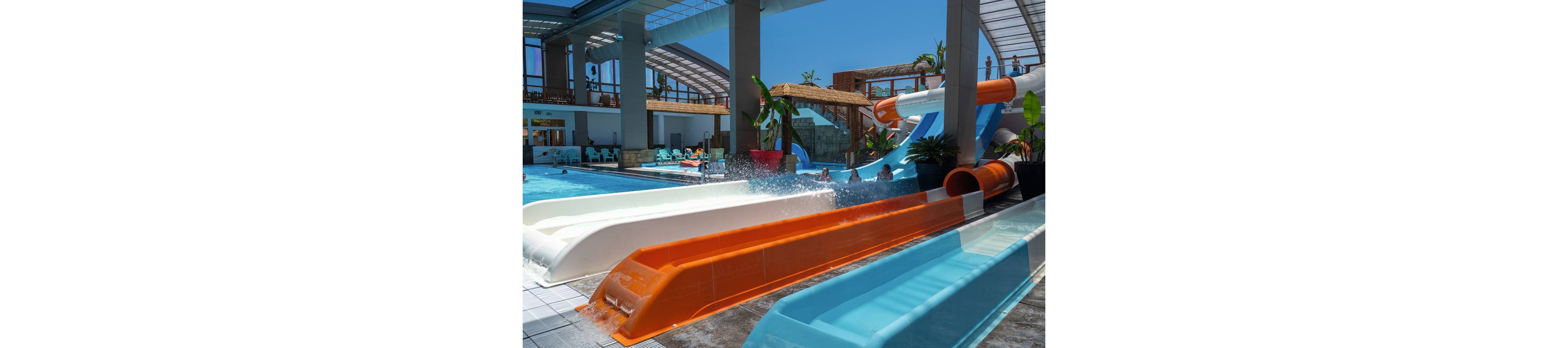 A set of brightly colored water slides empties into a large indoor pool area. People are splashing into the water from the slides while others relax in the background. The structure features high ceilings, glass panels, and tropical plants that create a resort-like atmosphere. Strong sunlight and blue skies are visible through the open sections, enhancing the vibrant, summery feel.