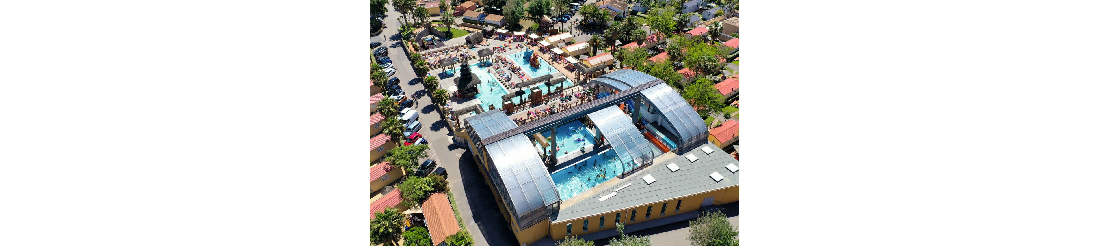An aerial view shows a large resort complex with multiple outdoor and covered swimming pools. Families and children enjoy the water, slides, and sun loungers around the pool deck. The surrounding area features rows of small vacation cabins and palm trees, creating a dense holiday village atmosphere. Bright colors, clear blue water, and sunny weather convey a lively summer vacation mood.