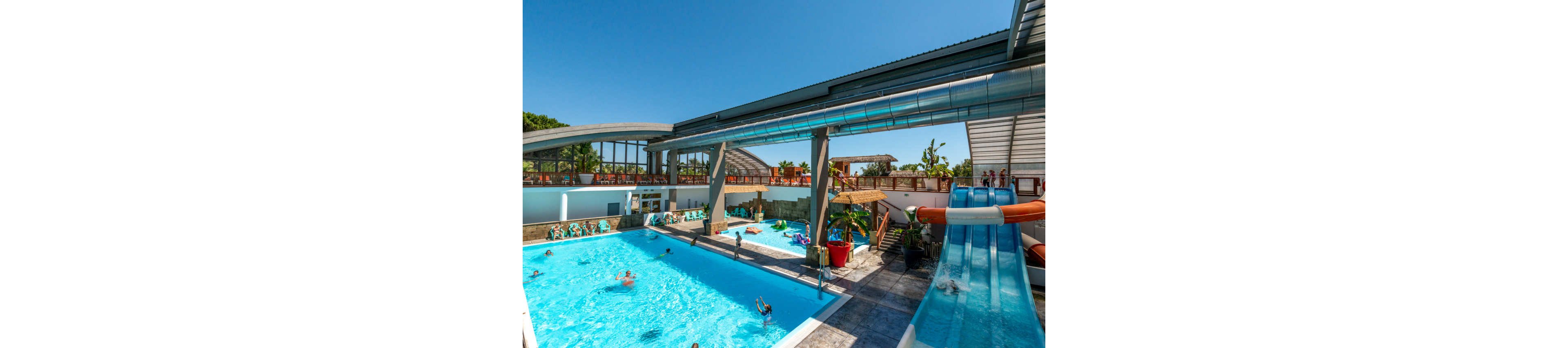 A bright outdoor swimming pool is filled with people enjoying the water under a clear blue sky. Large water slides curve down into the pool area, with one slide enclosed in a metal tube overhead. Sun loungers and umbrellas line the deck where visitors relax and watch the swimmers. A partially covered structure and elevated walkway add architectural interest to the modern aquatic center. The overall scene feels like a lively summer day at a family-friendly water park.