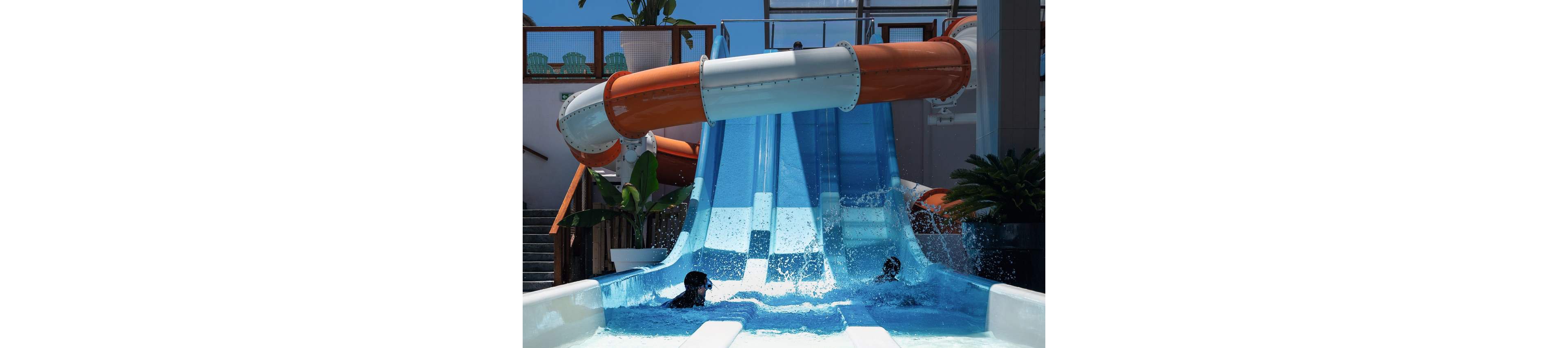 Two children splash into a pool at the bottom of bright blue parallel water slides. The slides descend from an enclosed orange and white tube structure in an outdoor water park. Sunlight highlights the flowing water and creates a playful, energetic scene. Tropical plants and seating areas are visible in the background, enhancing the vacation atmosphere.