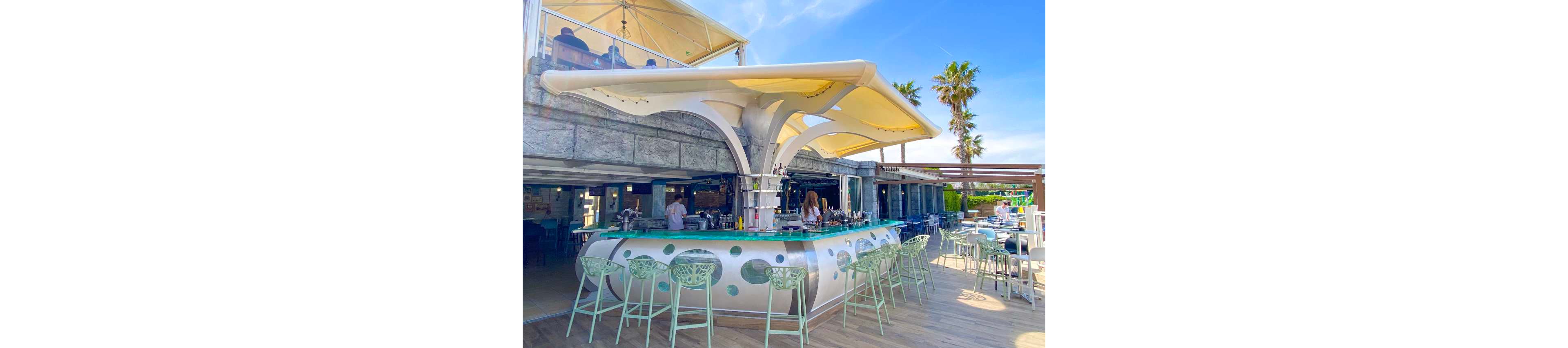 A contemporary open-air bar with a futuristic white canopy stands at the center of a coastal venue. Patrons sit and stand around the circular counter while bartenders prepare drinks. Light green bar stools and turquoise accents complement the bright, sunny seaside setting. Palm trees, blue sky, and outdoor seating create a relaxed vacation atmosphere.