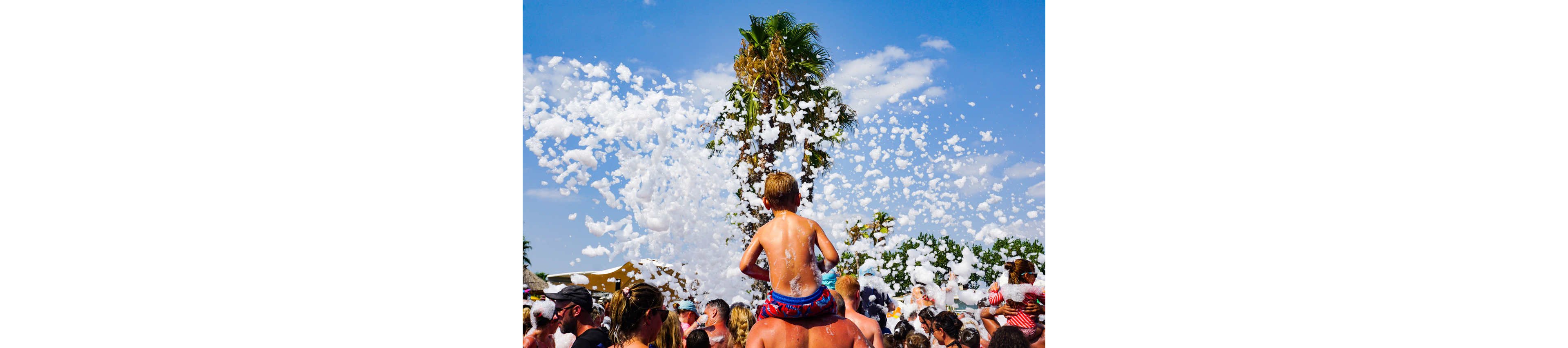 A young child sits on an adult’s shoulders in the middle of a crowded outdoor foam party. People are gathered closely together under a bright blue sky with a tall palm tree in the background. White foam fills the air, creating a playful and festive atmosphere. The scene suggests a warm, sunny day at a lively summer event or festival.