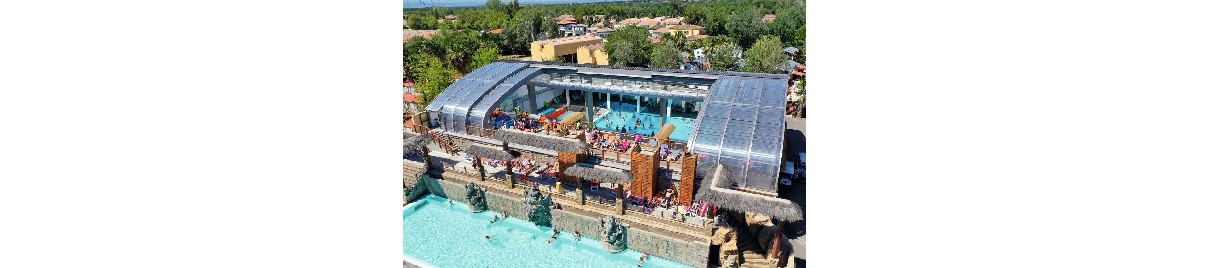 An aerial view shows a large resort complex with multiple swimming pools and a partially covered aquatic center. People relax on lounge chairs and swim in both the open-air and indoor pools. The structure features transparent curved roofing, wooden accents, and thatched details, giving it a tropical theme. Surrounding the water park are trees and low-rise buildings, suggesting a vacation destination in a warm climate.