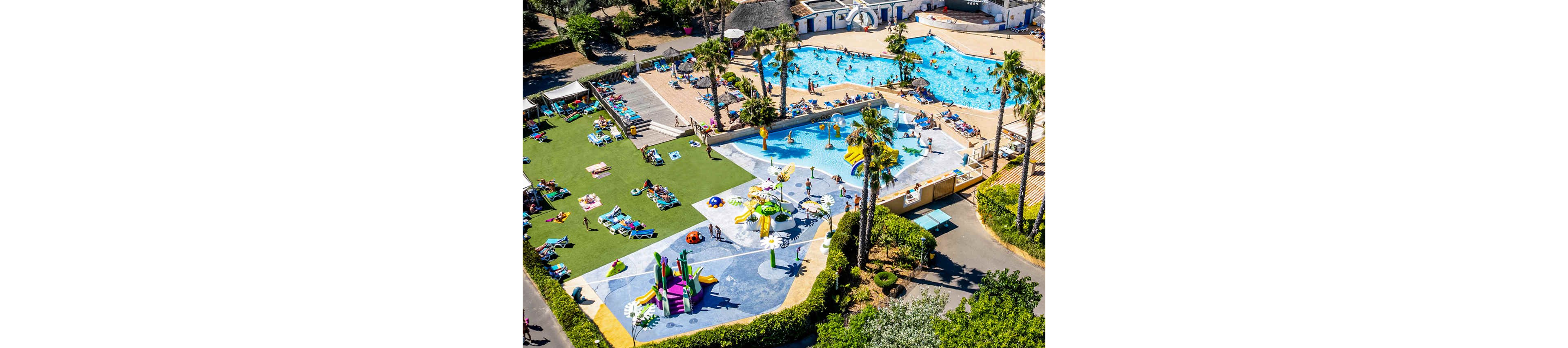 An aerial view shows a lively outdoor water park filled with families enjoying multiple pools and play areas. Children splash around colorful water slides and fountains while adults relax on lounge chairs surrounding the pools. Palm trees, green lawns, and bright umbrellas create a tropical resort atmosphere. The scene captures a sunny vacation day with people swimming, sunbathing, and playing in the water.