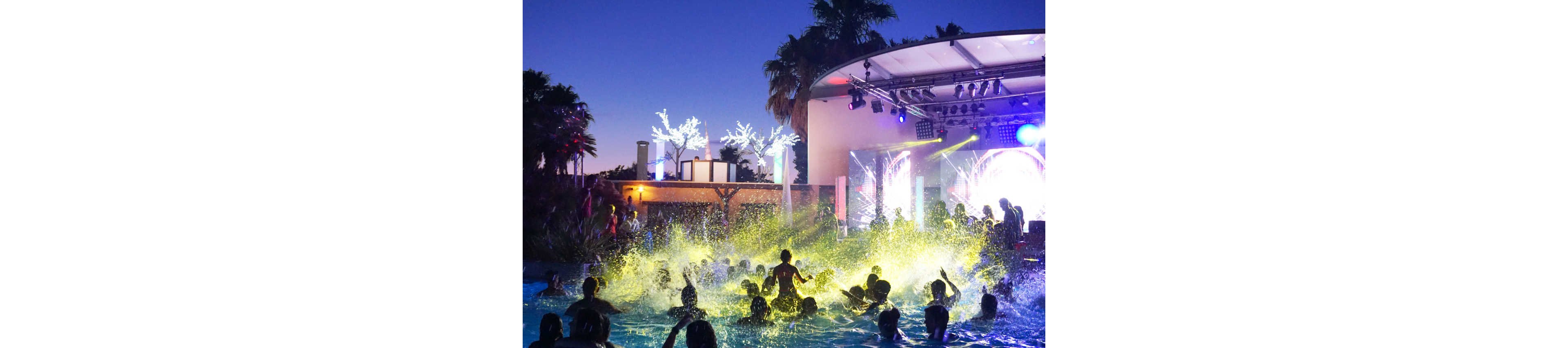 A lively crowd splashes in a swimming pool during a nighttime party. A stage with bright, colorful lights and screens illuminates the scene in front of the water. Palm trees and decorative lighted trees frame the outdoor venue under a clear evening sky. The image captures energetic movement, music, and a festive summer atmosphere.