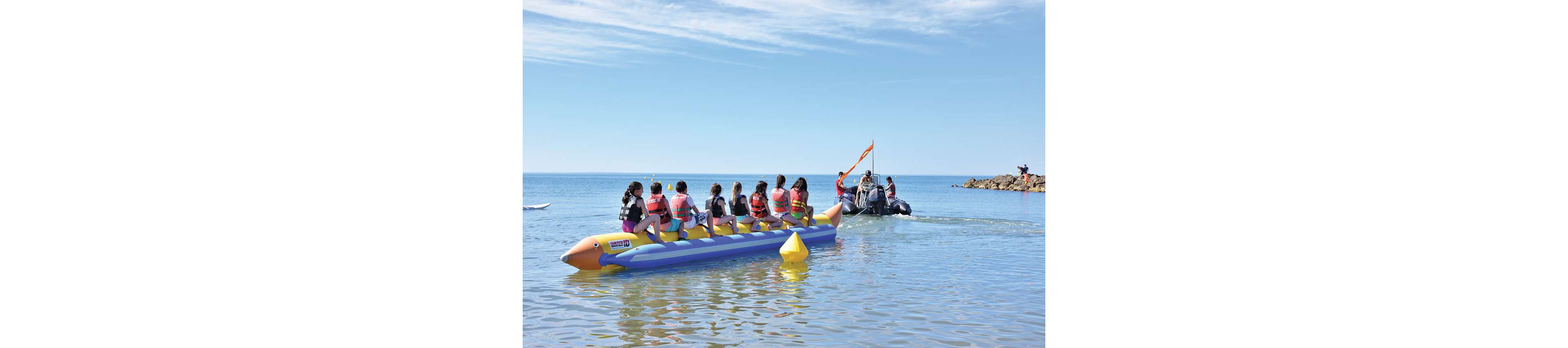 A group of people wearing life jackets ride an inflatable banana boat in shallow coastal water. They sit in a line facing forward as the boat is prepared to be towed by a motorboat ahead. The scene takes place on a calm, sunny day with clear blue sky and gentle waves. A yellow buoy floats nearby, and a rocky breakwater is visible in the distance.
