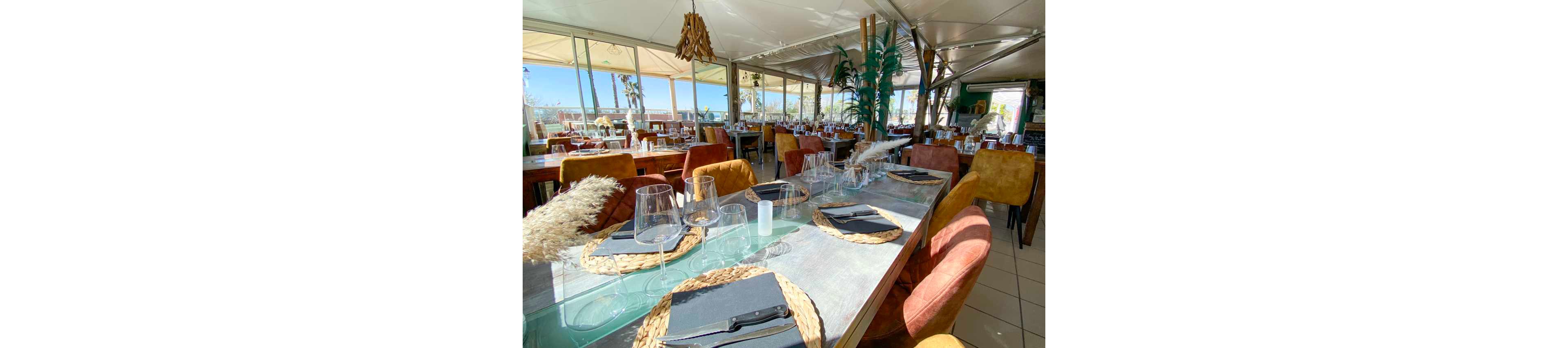 A bright restaurant interior shows a long table elegantly set for guests. Sunlight streams through large windows, revealing an outdoor terrace and palm trees in the background. Each place setting includes woven placemats, black napkins, cutlery, and multiple clear wine glasses. Warm-toned chairs and decorative plants create a relaxed, coastal dining atmosphere.