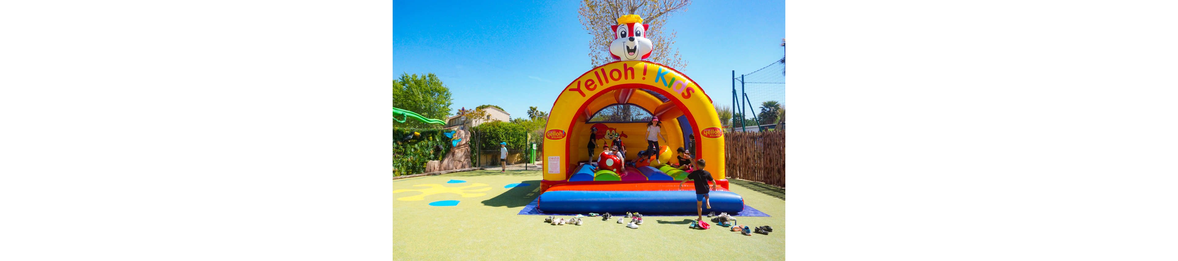 A group of children play on a bright inflatable bouncer in an outdoor playground. The yellow and red structure features a cartoon chipmunk character and the text "Yelloh! Kids" across the top. Shoes are scattered on the ground in front while kids jump and climb inside the bouncer. The scene is set on a sunny day with blue sky, trees, and a fenced area in the background.