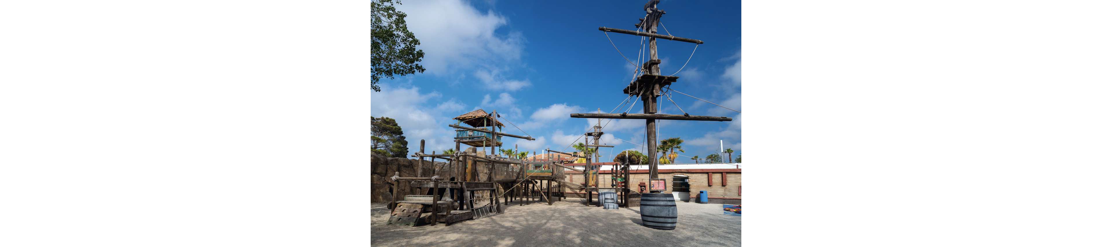 A large wooden pirate ship playground stands in an open outdoor area under a bright blue sky with scattered clouds. The structure features masts, ropes, platforms, and ladders, evoking a nautical adventure theme. Surrounding elements include barrels, fences, and nearby trees and palm trees, suggesting a themed park or recreational space. The scene is empty of people, highlighting the detailed construction and weathered wood textures.