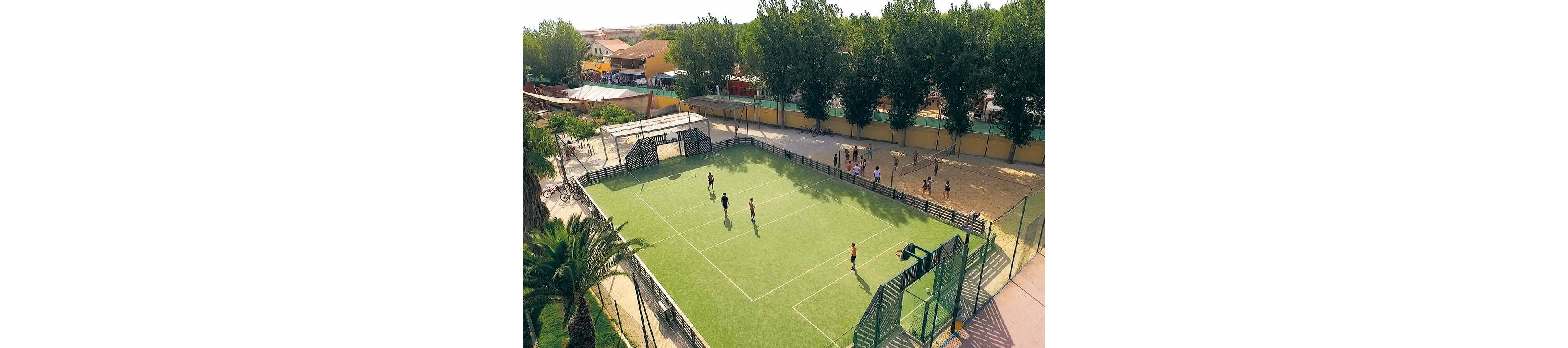 An aerial view shows a small outdoor sports court with a green artificial turf surface. A few people are scattered across the field, appearing to play or practice on a sunny day. The court is surrounded by high fences, nearby trees, and a residential neighborhood in the background. On one side, more people gather along a sandy area next to a long yellow wall.