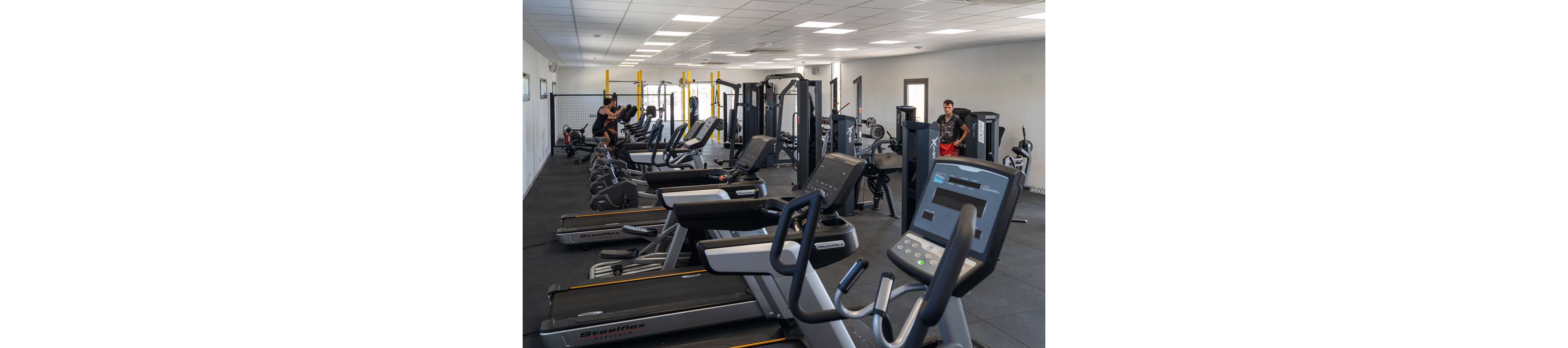 A spacious modern gym features rows of treadmills and various strength machines arranged neatly in a bright room. The setting includes a clean, open layout with large windows and a light ceiling, creating an airy atmosphere. A few people are exercising on different machines, adding a sense of activity and energy. The overall palette is neutral with black flooring and grey fitness equipment, emphasizing a professional workout environment.