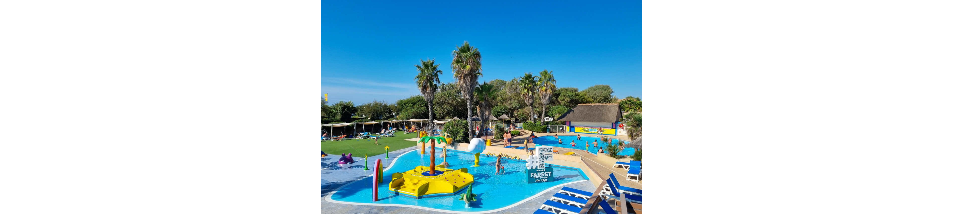 A vibrant outdoor swimming pool features children and adults playing in bright blue water. The pool includes playful structures such as a yellow floating platform with palm tree decorations and a small slide. Palm trees, green lawns, and sun loungers surround the pool area under a clear blue sky. Families relax and play throughout the resort-style setting, creating a lively vacation atmosphere.