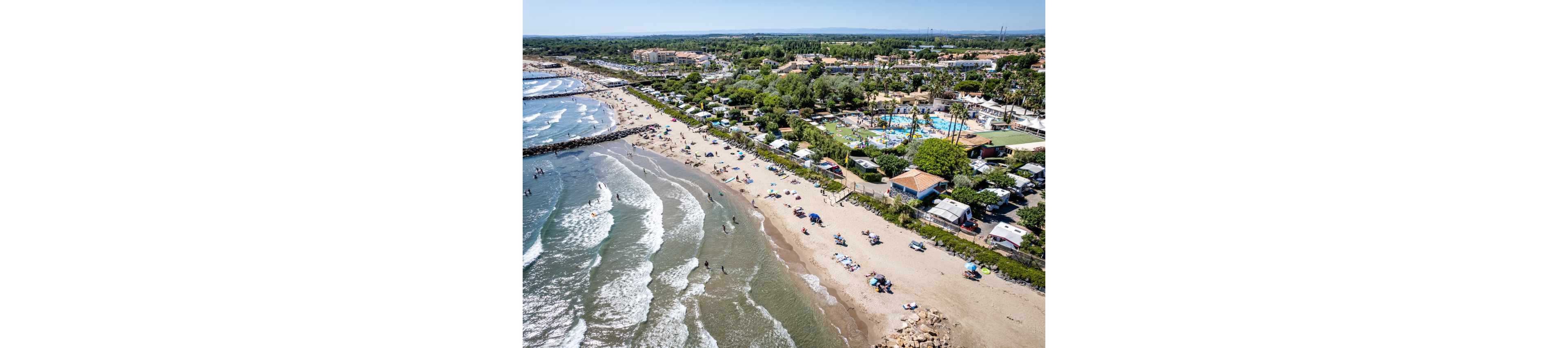 An aerial view shows a long sandy beach lined with sunbathers and umbrellas beside gentle ocean waves. People are swimming, surfing, and relaxing along the shoreline while others enjoy the water near breakwaters. Adjacent to the beach, a large campground with rows of caravans, tents, and bungalows is visible among trees. A bright blue swimming pool complex and surrounding resort facilities stand out in the middle of the campsite. In the distance, flat coastal plains and low hills stretch toward the horizon under a clear sky.