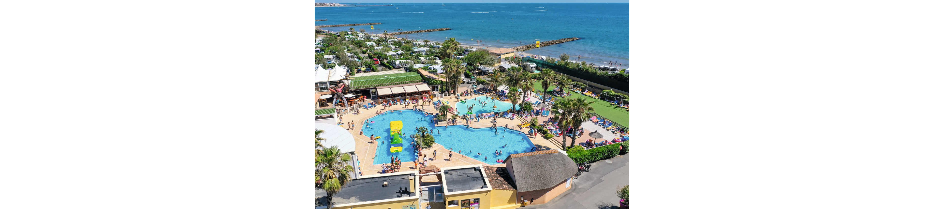 An aerial view shows a busy resort swimming pool complex near the sea. Families and children enjoy multiple pools, slides, and sun loungers surrounded by palm trees. The resort buildings and landscaped areas frame the water park, while the sandy beach and calm ocean stretch out in the background. The bright colors and clear weather create a lively, summery vacation atmosphere.