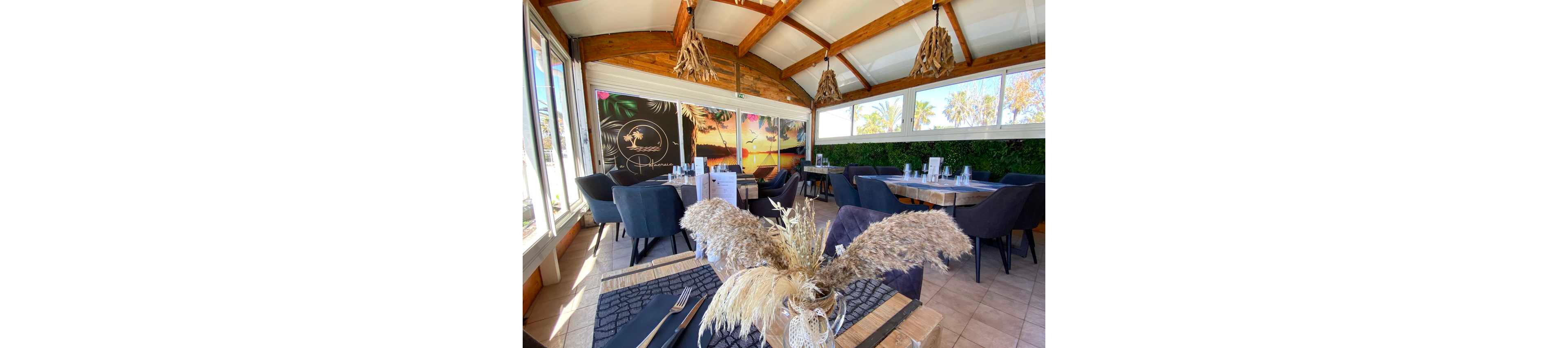 A stylish restaurant dining area features wooden tables set with dark placemats and neatly arranged glassware. Large windows let in abundant natural light, revealing palm trees and a sunny outdoor view. Soft upholstered chairs surround the tables, while dried pampas grass centerpieces add a natural decorative touch. Warm wooden ceiling beams and hanging light fixtures contribute to a cozy yet contemporary atmosphere.