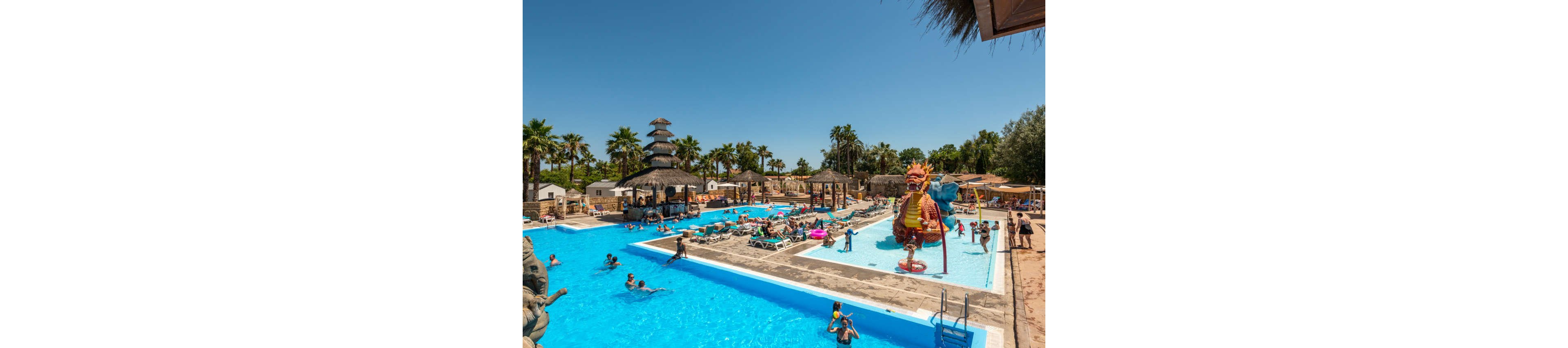 A large outdoor resort pool is filled with families enjoying a sunny day. Children play around a colorful dragon-themed water playground while adults relax on sun loungers. Palm trees, thatched umbrellas, and cabana-style structures surround the pool area, creating a tropical atmosphere. The bright blue water and clear sky emphasize a warm, summer vacation setting.