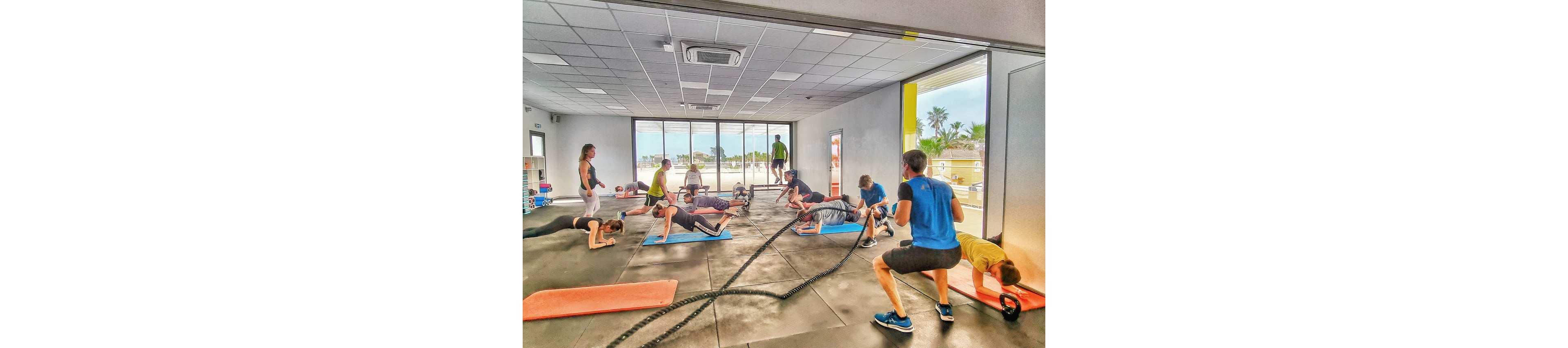 A group of adults participates in an indoor fitness class using battle ropes and floor mats. The instructor guides the session while participants perform planks, squats, and other strength exercises. Large windows reveal an outdoor view with palm trees and bright daylight. The scene captures an energetic workout atmosphere in a modern gym studio.