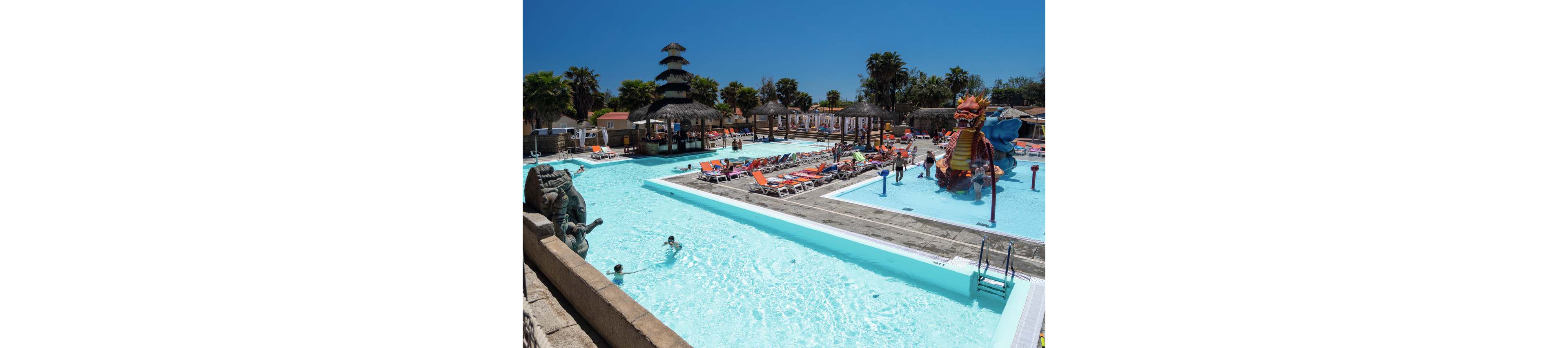 A large outdoor resort pool features a colorful dragon-themed water slide and shallow swimming areas. Families and children enjoy the bright blue water under a clear sunny sky. Rows of orange sun loungers and umbrellas line the poolside, with palm trees and thatched structures in the background. The scene conveys a fun, tropical vacation atmosphere at a family-friendly water park.