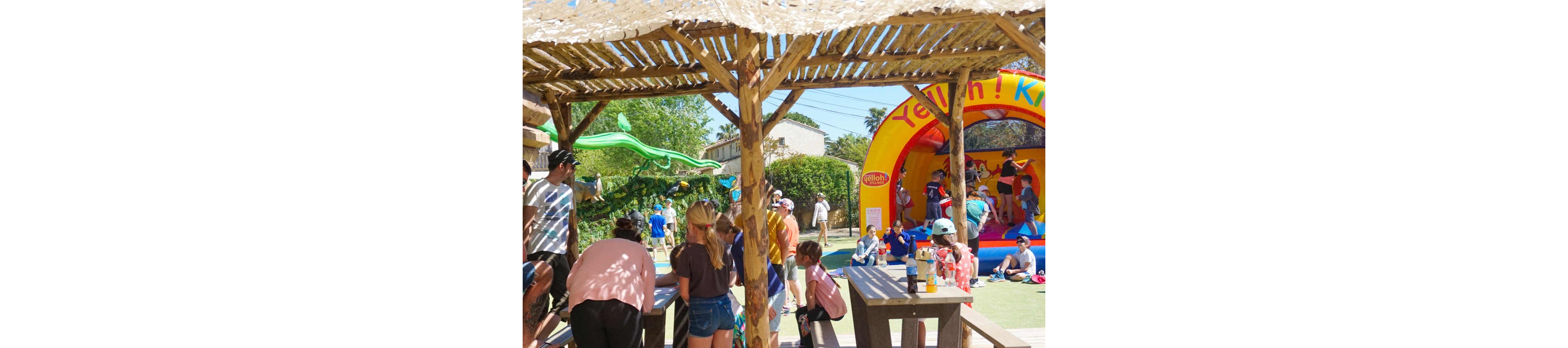 A group of children and adults gather under a wooden shade structure at an outdoor play area. In the background, kids jump and play inside a colorful inflatable bouncy castle. Other visitors relax nearby while children explore the playground and slide. The bright sunlight and clear sky create a cheerful, summery atmosphere.