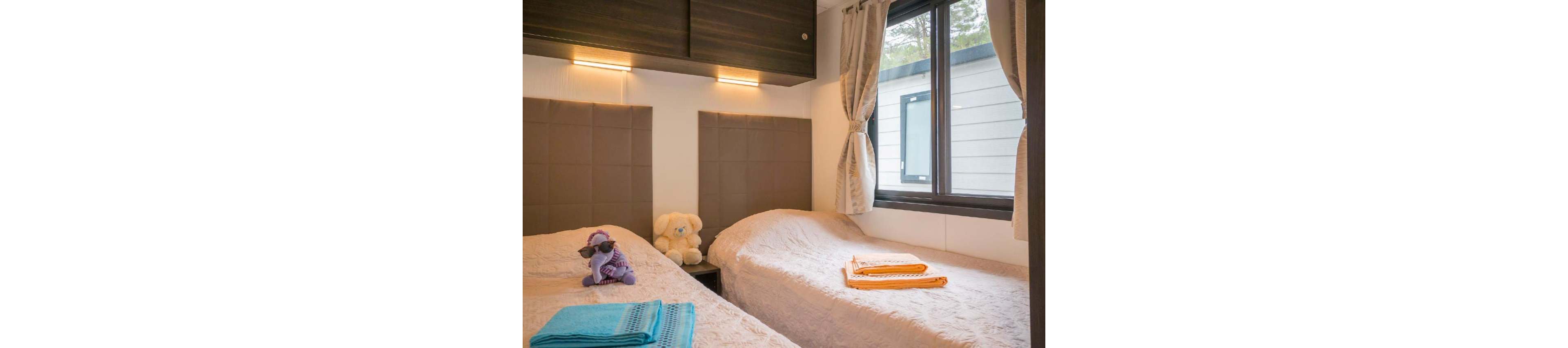 A compact bedroom features two single beds separated by a padded headboard wall. Soft lighting from above the headboards creates a warm and inviting atmosphere. A teddy bear and a small plush toy sit on the beds, adding a playful touch. Folded towels in bright colors are placed on each bed, and a large window with curtains lets in natural light and a view of the exterior.