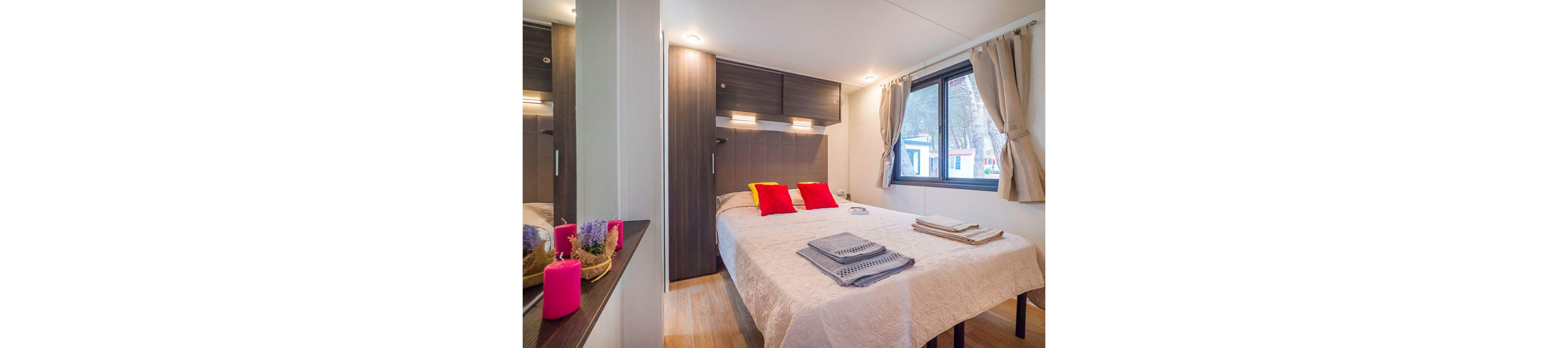 A bright modern bedroom features a double bed neatly made with a light quilt and colorful accent pillows. Folded towels and linens are arranged on the bed, suggesting a guest-ready space. Warm wood flooring, built-in dark wood cabinets, and soft wall lighting create a welcoming atmosphere. A large window with beige curtains lets in natural light and offers a view of neighboring structures, while decorative candles add a subtle touch of color.