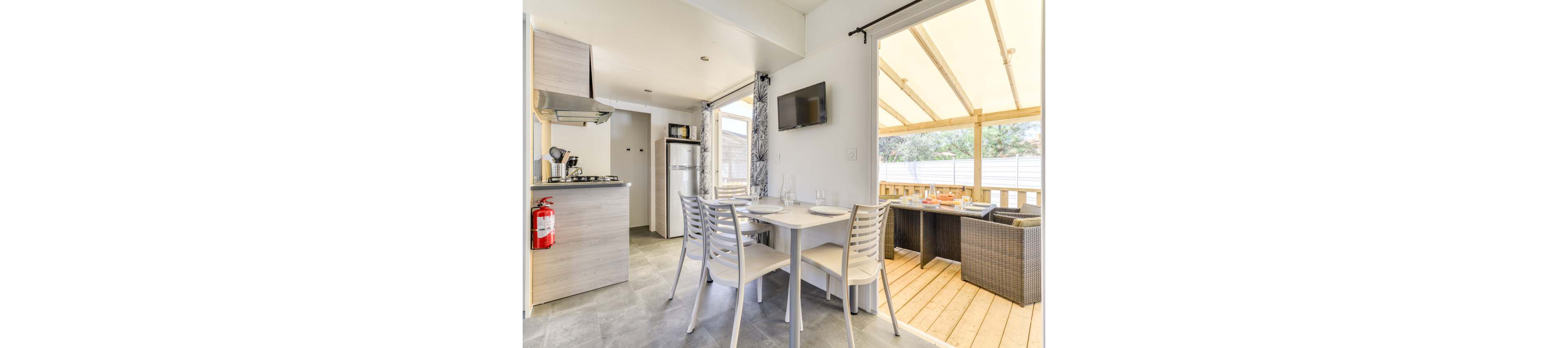 A compact modern kitchenette opens onto a bright covered patio dining area. Inside, a small dining table with four white chairs sits near a wall-mounted television and a simple galley kitchen with stove and refrigerator. Large windows and a sliding door bring in natural light and reveal an outdoor seating area with a wicker-style table and chairs on a wooden deck. Neutral tones and clean lines create a fresh, contemporary atmosphere suitable for vacation or small-apartment living.