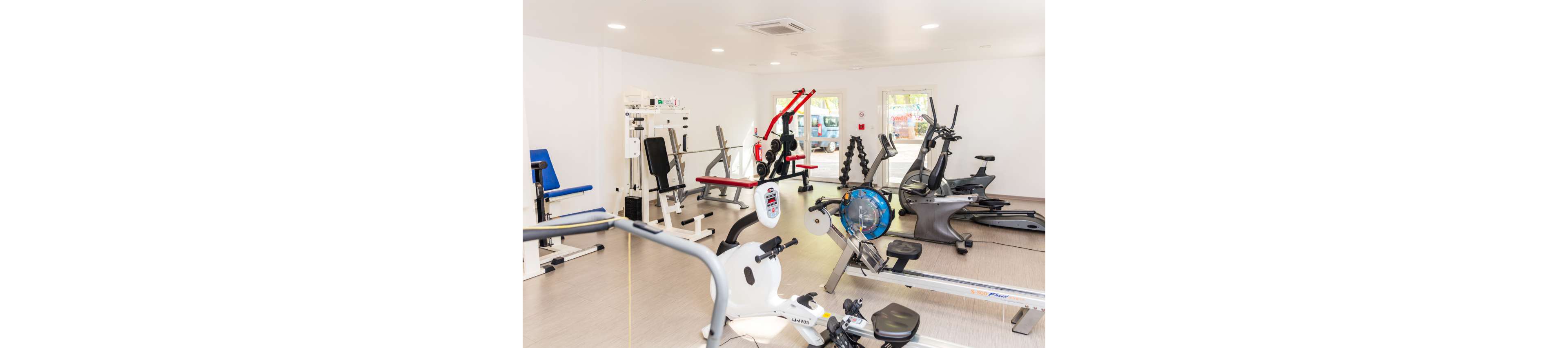 A bright, modern gym room features a variety of exercise machines neatly arranged. Cardio equipment such as stationary bikes and elliptical trainers stand alongside strength-training machines and benches. Large windows allow natural light to fill the clean, white space, creating an inviting workout environment. The room appears organized and spacious, suitable for individual fitness routines.