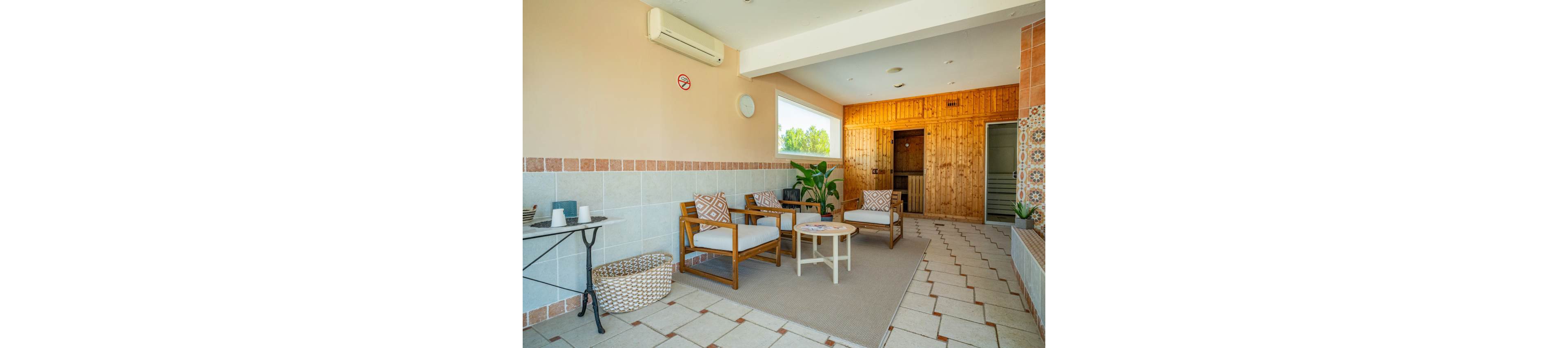 A cozy indoor lounge features wooden armchairs with patterned cushions arranged around a small round coffee table. The space includes tiled floors and walls, a woven basket, and a side table with candles and cups. Large windows bring in natural light and greenery from outside, while a wooden-paneled wall at the back leads to a sauna or relaxation room. Neutral tones and soft textures create a calm, spa-like atmosphere.