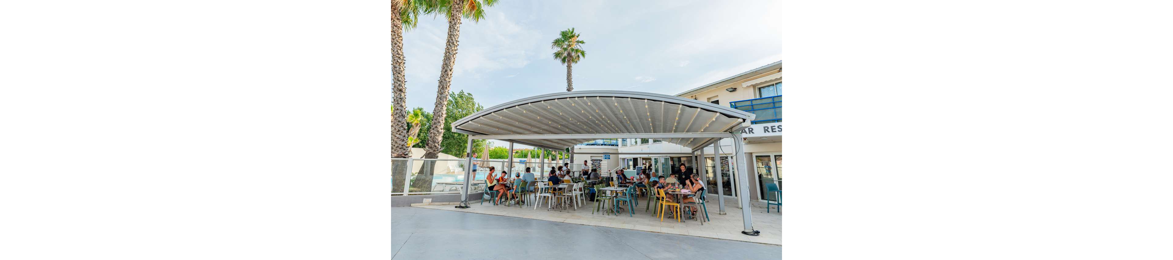 An open-air dining area at a resort features guests seated under a large curved canopy. People sit at colorful tables and chairs, enjoying food and drinks in a relaxed atmosphere. The terrace is adjacent to a modern building with balconies and a visible pool area behind glass fencing. Tall palm trees and clear skies emphasize the warm, sunny climate. String lights under the canopy add a festive touch to the scene.